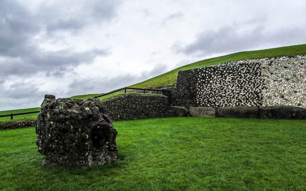 Slope of Newgrange against a rainy sky. Foreground shows a large loose piece separated from main structure.